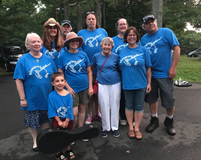 a group of people stand together smiling, all wearing blue shirts wiht the Bluegrass Festival logo in white. a small boy at the front holds a guitar case.