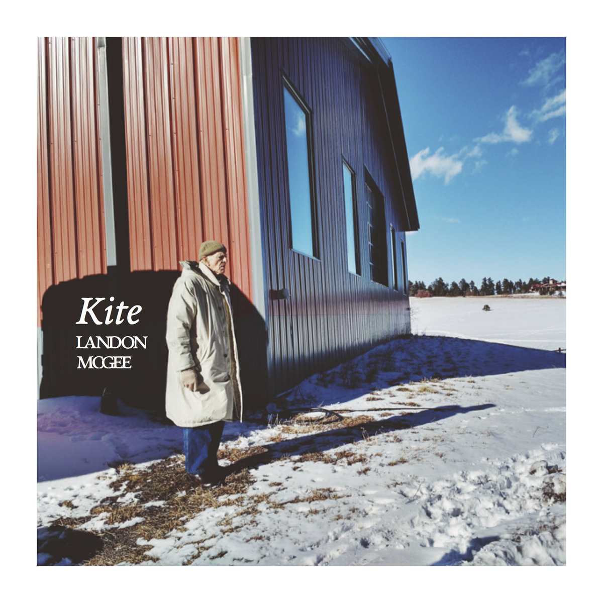 sunny, blue-toned shot of a red barn in the snow, with an olderm an standing apart and looking to the side, just out of the barn's stark shadow, wearing a functional and sharp winter coat, hat, and gloves in tones of grey, and blue jeans.